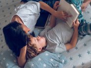 Von oben Aufnahme eines süßen Jungen, der auf einem schönen Bett liegt und mit Hilfe seiner Mutter ein interessantes Buch liest — Stockfoto