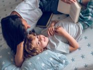 Von oben Aufnahme eines süßen Jungen, der auf einem schönen Bett liegt und mit Hilfe seiner Mutter ein interessantes Buch liest — Stockfoto