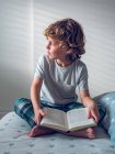 Garçon mignon en pyjama assis sur un lit confortable et la lecture beau livre — Photo de stock
