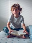 Симпатичный мальчик в пижаме сидит на удобной кровати и читает хорошую книгу — стоковое фото