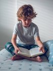 Симпатичний хлопчик у піжамі сидить на зручному ліжку і читає гарну книгу — стокове фото