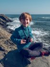 Fröhlicher kleiner Junge sitzt mit Fernglas auf Stein am Meer — Stockfoto