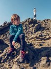 Young boy sitting on rocky hill on background of beacon tower — Stock Photo