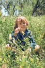 Портрет улыбающегося мальчика, сидящего в траве и полевых цветах и держащего цветок — стоковое фото