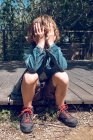 Little boy sitting on wooden footpath in countryside with hands over face — Stock Photo