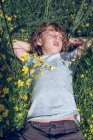 Mignon petit garçon rêvant avec les yeux fermés tout en étant couché sur l'herbe haute avec des fleurs colorées — Photo de stock