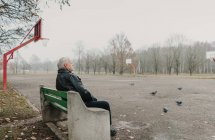Senior man in leather jacket sitting on bench near sports ground in park and looking at pigeons — Stock Photo