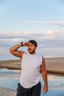 Взрослый мужчина с бородой в спортивной одежде, держась за руку у лба и глядя на расстояние, стоя на пляже во время тренировок на открытом воздухе — стоковое фото