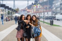 Atractivas señoras alegres tomando selfie en el teléfono móvil y caminando por sendero cerca de edificios antiguos en Oporto, Portugal - foto de stock