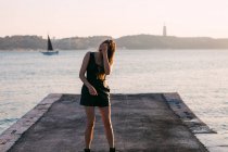 Улыбающаяся очаровательная молодая женщина в черной одежде и сапогах позирует на набережной рядом с поверхностью воды с яхтой на закате — стоковое фото