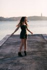 Sonhador encantador jovem mulher em preto desgaste e botas dançando em aterro perto da superfície da água ao pôr do sol — Fotografia de Stock