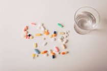 Різнокольорові таблетки та капсули, розкидані склянкою води на білому тлі — стокове фото