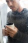 Tiro através de vidro janela molhada do homem em pé suéter com xícara de café — Fotografia de Stock