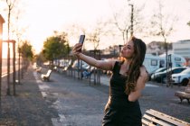 Senhora atraente em vestido preto com mão no cabelo tomando selfie na rua ao pôr do sol — Fotografia de Stock