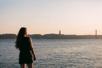 Giovane signora in abito nero in piedi su argine vicino alla superficie dell'acqua al tramonto e sognando — Foto stock