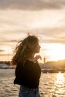 Attrayant jeune femme avec les cheveux bouclés regardant loin tout en se tenant près de l'eau pendant le coucher du soleil en ville — Photo de stock