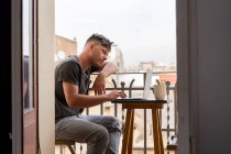 Side view of casual adult man sitting at small table on balcony using laptop and having cup of coffee — Stock Photo