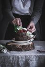 Женские руки украшают шоколадный торт, украшенный малиной и цветами на тарелке на деревянном стенде — стоковое фото