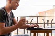 Beau homme avec une tasse de café et en utilisant un ordinateur portable sur le balcon — Photo de stock