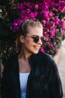 Lovely young lady in trendy outfit and sunglasses looking away while standing near bush with beautiful flowers on street — Stock Photo