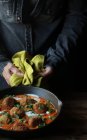 Человеческие руки держат тяжелую сковородку вкусной цветной капусты и шары из киноа с соусом и петрушкой над деревянным столом — стоковое фото