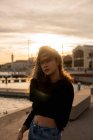 Schöne junge Frau mit Haaren im Gesicht, die bei Sonnenuntergang am Ufer der Stadt steht — Stockfoto
