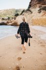 Vista trasera de la joven descalza con cámara fotográfica caminando sobre la arena cerca del agua del mar - foto de stock
