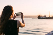 Vue arrière du bateau de tir femme sur smartphone sur le remblai près de l'eau au coucher du soleil — Photo de stock