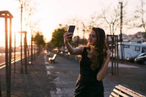 Senhora atraente em vestido preto com mão no cabelo tomando selfie na rua ao pôr do sol — Fotografia de Stock
