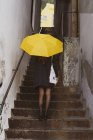 Vista posterior señora delgada en abrigo con paraguas amarillo y de pie en las escaleras entre las paredes de Oporto, Portugal - foto de stock