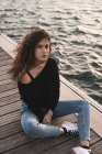 Молода жінка дивиться на камеру, сидячи на дерев'яному пірсі біля води — стокове фото
