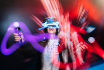 Borroso tiro de larga exposición de la mujer feliz en gafas VR jugando videojuego con controladores en la oscuridad - foto de stock