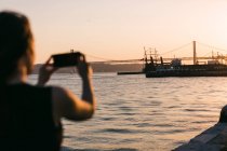 Vue arrière du bateau de tir femme sur smartphone sur le remblai près de l'eau au coucher du soleil — Photo de stock
