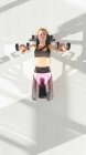 Von oben junge schlanke Dame in Sportbekleidung auf Bankdrücken liegend und Brusttraining mit Kurzhanteln im Fitnessstudio auf grauem Hintergrund — Stockfoto