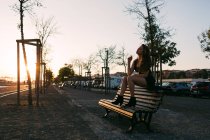 Sensual jovem senhora em vestido preto sentado no banco na rua no beco ao pôr do sol — Fotografia de Stock