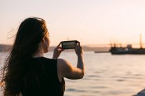 Vista trasera del barco de tiro de mujer en el teléfono inteligente en el terraplén cerca del agua al atardecer - foto de stock