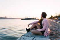 Мечтательная чувственная юная леди, сидящая на набережной возле поверхности воды на закате — стоковое фото