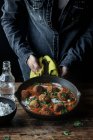 Человеческие руки держат тяжелую сковородку вкусной цветной капусты и шары из киноа с соусом и петрушкой над деревянным столом — стоковое фото