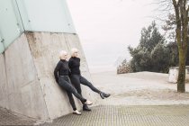 Mulheres jovens atraentes em desgaste escuro esticando as pernas e inclinando-se na parede na rua perto de árvores na Lituânia — Fotografia de Stock
