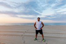 Homem bonito positivo em sportswear mantendo as mãos na cintura enquanto estava em pé na praia de areia durante o pôr do sol — Fotografia de Stock