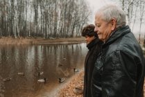 Вид сбоку на пожилых мужчину и женщину, стоящих у воды и смотрящих на уток в осеннем парке — стоковое фото