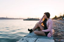 Sognante sensuale giovane signora seduta su argine vicino alla superficie dell'acqua al tramonto — Foto stock