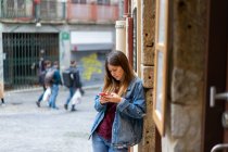 Mujer atractiva en chaqueta de jean sosteniendo el teléfono móvil rojo y de pie cerca de la pared en la calle en Oporto, Portugal - foto de stock