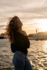 Приваблива молода леді з кучерявим волоссям, торкаючись плеча і дивлячись на камеру, стоячи біля води під час заходу сонця в місті — стокове фото