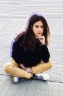 Чарівна молода жінка в модному вбранні дивиться на камеру, сидячи на тротуарі на міській вулиці — стокове фото