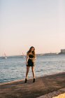 Giovane signora in abito nero e stivali in posa su argine vicino alla superficie dell'acqua con yacht nella giornata di sole — Foto stock