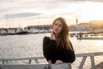 Portrait of young lady touching curly hair and looking at camera while leaning on railing on blurred background of pier and water — Stock Photo