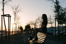 Чувственная юная леди в черном платье сидит на скамейке на улице в переулке на закате — стоковое фото