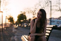 Porträt einer jungen Frau im schwarzen Kleid, die bei Sonnenuntergang auf einer Bank auf der Straße sitzt — Stockfoto