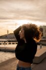 Привлекательная юная леди с кудрявыми волосами, смотрящая в сторону, стоя возле воды во время заката в городе — стоковое фото